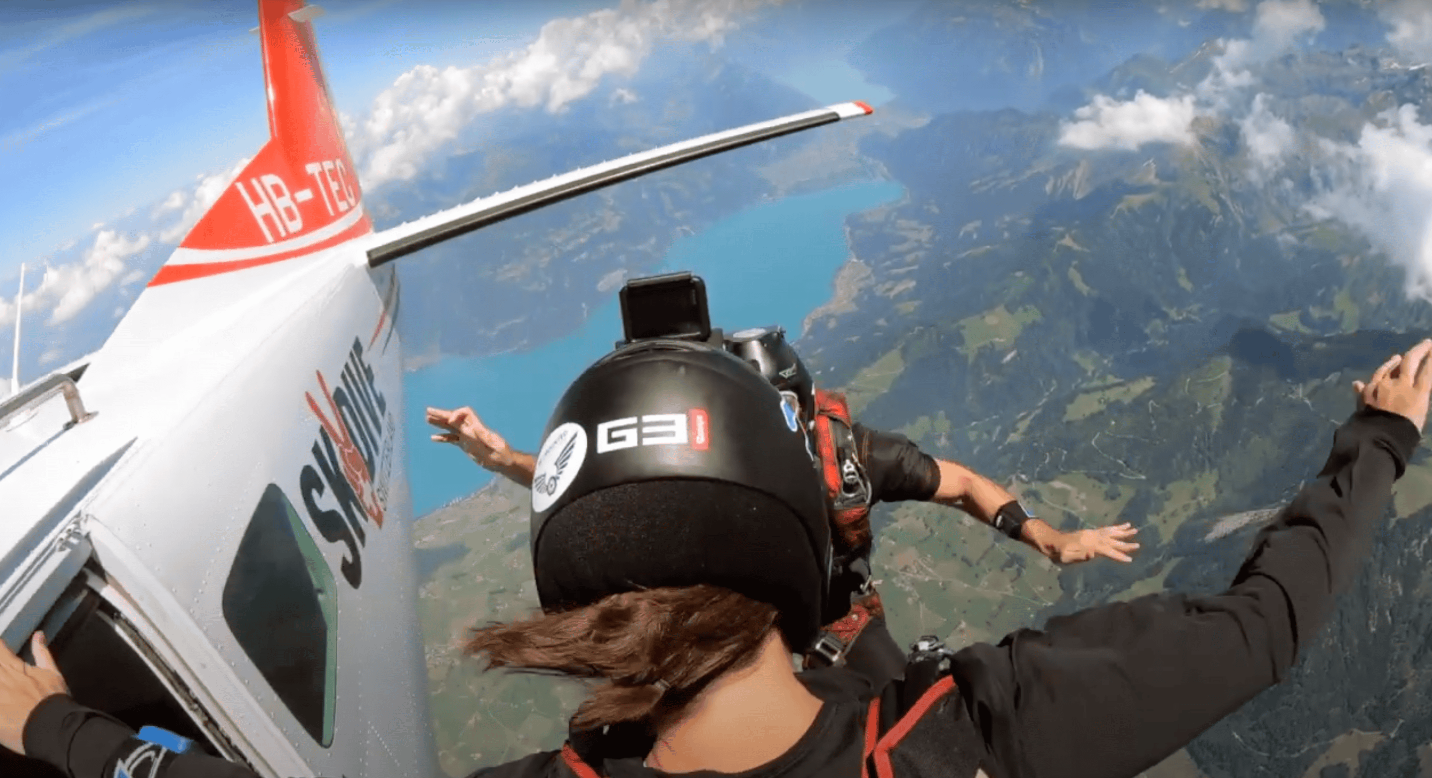 A girl and a guy jumping out of a plane in Interlaken for a skydiving activity. It is a beautiful day with the lake in the background.